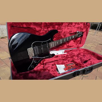 Černá elektrická kytara s tvarem typu Stratocaster v otevřeném kufru s červeným vnitřkem. Na hlavě kytary je bílý nápis Ibanez. Kytara má chromovaný…