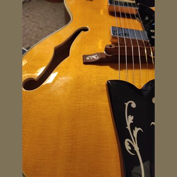 Detailní pohled na tělo žluté polodupaté elektrické kytary s f-otvorem. Na dřevěném kobylníku je viditelné logo znacky Epiphone. Kytara má dva chromo…
