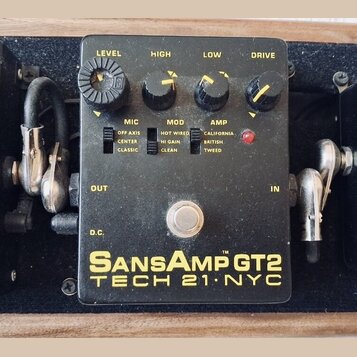 Černý efekt pedál s nápisy SansAmp GT2 a TECH 21 NYC. Na horní straně jsou čtyři ovládací knoflíky s popisky LEVEL, HIGH, LOW a DRIVE. Pod nimi se na…