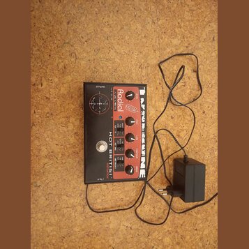 Kytarový efekt pedál s černým povrchem a červenými ovládacími knoflíky. Na zařízení jsou viditelné nápisy RADIAL, HOT BRITISH DISTORTION a LINE 6. V…