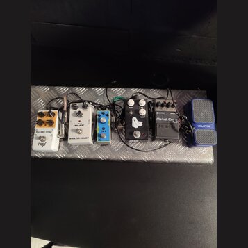Sestava efektů na stříbrném kovovém pedalboardu s protiskluzovou strukturou. Zleva jsou vidět pedály s nápisy: „PHASER CORE“ a „nux“, „NUX ANALOG DEL…