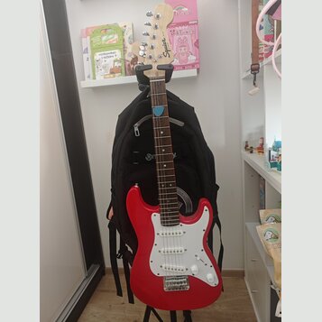 Červená elektrická kytara s bílým pickguardem a hnědým hmatníkem. Na hlavě nástroje je viditelné logo Squier. Kytara stojí na stojanu před černým bat…
