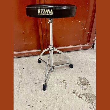 Černý bubenířský židle s kovovým chromovaným stojanem a třínožkou. Na sedáku je bílý nápis TAMA a pod ním text THE STRONGEST NAME IN DRUMS. Povrch se…