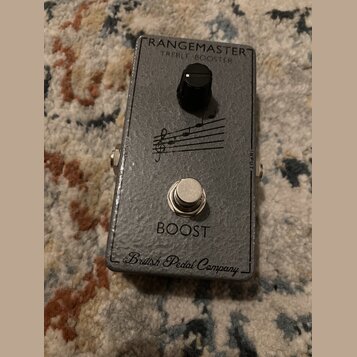 Šedý efekt pedál s texturovaným povrchem. Na horní části je nápis RANGEMASTER TREBLE BOOSTER, uprostřed je černý ovládací knoflík a grafika s hudební…
