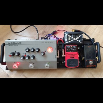 Sestava efektů na černém pedalboardu. Vlevo je stříbrný pedál s nápisy MODULE, TERRA-NOVA a BASS PREAMP PEDAL. Uprostřed je červený pedál s nápisy CO…