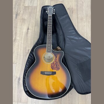 Akustická kytara s vyvedeným korpusem v provedení sunburst, položená v černém měkkém pouzdře. Na hlavě nástroje je bílý nápis GUILD. Kytara má hnědý…