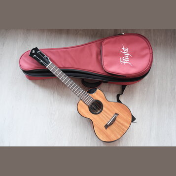 Dřevěný akustický nástroj typu ukulele v přirozené barvě s černým lemováním. Na hlavě hmatníku je bílé logo znázorňující stylizované písmeno S. Vedle…