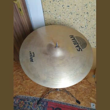Zlatavý kovový činel na stojanu. Na povrchu je černý nápis SABIAN s doprovodným logem a text AA Heavy Ride. Povrch vykazuje jemné koncentrické rýhy a…