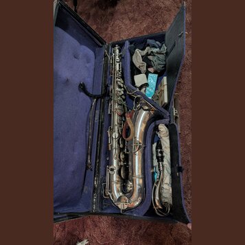 Stříbrný saxofon v otevřeném kufru s fialovým vnitřkem. K nástroji patří munzpun s mouthpiecem a samostatný hnědý řemínek. V kufru jsou rovněž umístě…