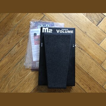 Černý volume pedál s textem M2 PASSIVE VOLUME. Na bocích jsou viditelné nápisy OUT a IN. Vedle pedálu je v plastovém sáčku dokumentace s nápisem MORL…