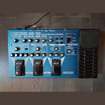 Modrý efektový pedál Boss ME-50 Guitar Multiple Effects. Na horní straně jsou různé ovládací knofíky, přepínače a červené digitální displeje. V dolní…