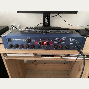 Modrý kytarový procesor s kovovým šasi a názvem V-AMP PRO a logem BEHRINGER. Na předním panelu jsou ovládací knofíky, červené tlačítka A až E a digit…