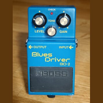 Modrý kytarový efekt s nápisy Blues Driver a BD-2. Na horní části jsou tři ovládací knoflíky s popisky LEVEL, TONE a GAIN a červená kontrolka CHECK.…