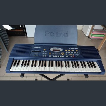 Modrý elektronický klavír umístěný na černém stojanu. Na horním panelu je nápis Roland a označení modelu EM-20. Nástroj má bílé a černé klávesy, řadu…