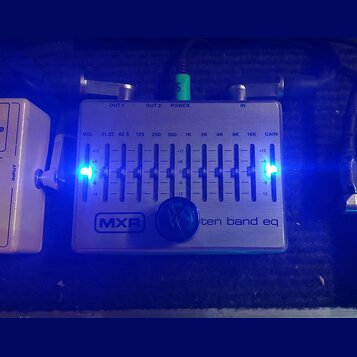 Stříbrný kytarový pedál s nápisy MXR a ten band eq. Zařízení obsahuje deset posuvníků pro ekvalizaci s označenmi frekvencí od 31.25 do 16K a dva posu…