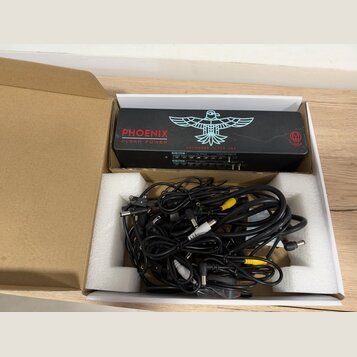 Černý napájecí adaptér s nápisy PHOENIX CLEAN POWER a HANDMADE IN THE USA, doplněný bílým logem ptáka. V krabici s polystyrenovými vložkami se nacház…
