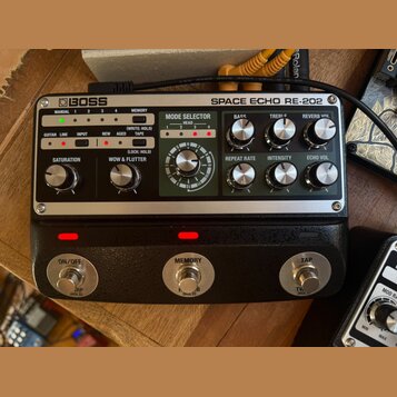 Stříbrný efektový pedál s černým spodním tělem a nápisy BOSS SPACE ECHO RE-202. Horní panel obsahuje ovládací knoflíky, přepínače a sekci MODE SELECT…