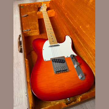 Elektrická kytara s červeno-oranžovým gradientem (sunburst) v hnědém kufru s oranžovou výplní. Nástroj má bílý pickguard, javorový hmatník a chromova…
