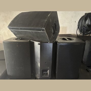Tři černé reproduktory s kovovými mřížkami. Jeden je umístěn v horní části jako monitor, dva další stojí vedle sebe pod ním. Povrchy jsou matně černé…
