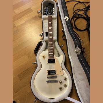 Kytara s bílým trupem a hnědým hmatníkem v černé kostelní kytarové skříni. Na hlavě je viditelné logo Gibson. Nástroj má dva humbuckery, čtyři otočné…