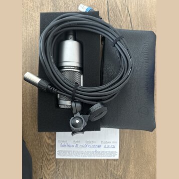 Stříbrný kondenzátorový mikrofon s mřížkou, na kterém je nápis audio-technica. Součástí je černý kabel s XLR konektory, černý držák a černý ochranný…