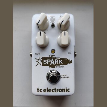 Bílý kytarový efekt s čtyřmi bílými oválnými knoflíky. Na pedálu jsou nápisy FAT CLEAN MID, LEVEL, TREBLE a logo SPARK BOOSTER s motivem zapálené záp…
