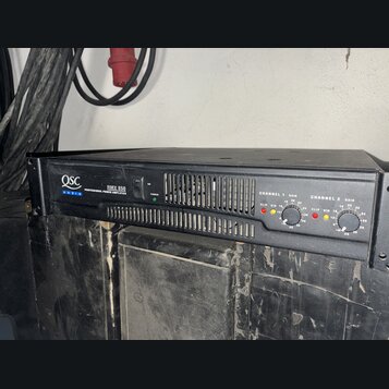 Černý výkonový zesilovač s nápisy QSC AUDIO a RMX 850 PROFESSIONAL POWER AMPLIFIER. Na předním panelu jsou dvě sekce označené CHANNEL 1 a CHANNEL 2,…