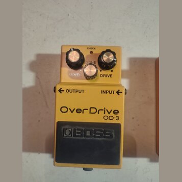 Žlutý efekt pedál s černým spínacím tlačítkem. Na horní části jsou tři ovládací knoflíky s nápisy LEVEL, TONE a DRIVE, nad nimi je nápis CHECK a červ…