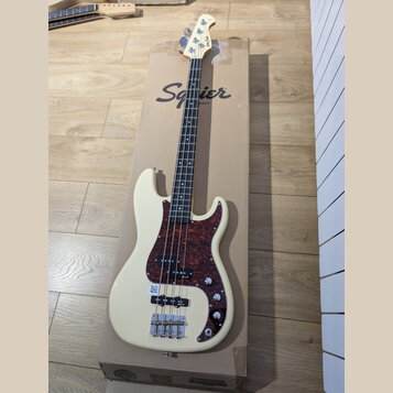 Kremově bílá elektrická basová kytara s hnědo-černým želvovinovým pickguardem, položená na kartonu s nápisem Squier by Fender. Na hlavě hmatníku je v…