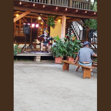 Fotografie zachicuje muzikanta na dřevěném pódiu, který hraje na akustickou kytaru světlé barvy. Vedle něj je umístěn mikrofon na stativu a černý rep…