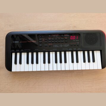 Černý přenosný digitální klavír s bílými a černými klávesami. Na levé straně je bílý nápis YAMAHA a model PSS-A50. Přístroj má ovládací panel s různý…