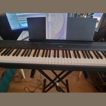 Černý digitální klavír značky YAMAHA umístěný na černém X-kovovém stojanu. Na klavíru je nápis YAMAHA a v zadní části je umístěna kniha s nápisem DIG…