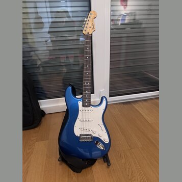 Modrá elektrická kytara s bílým pickguardem a javorovým hmatníkem. Na hlavě je viditelné logo Fender. Kytara stojí na černém stojanu a vedle ní je če…