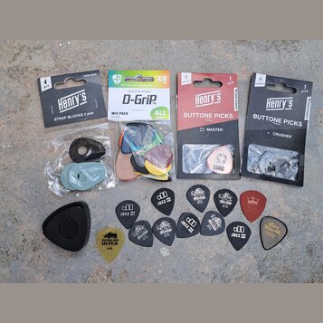 Fotografie zobrazuje sadu kytarového příslušenství. Jsou zde čtyři originální balení: „Henry's STRAP BLOCKS 2 mm“, „JANICKER PICKS D-Grip™ MIX PACK N…