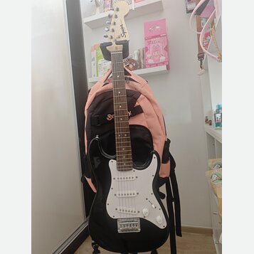 Černá elektrická kytara s bílým pickguardem a světle hnědým hmatníkem. Na hlavě nástroje je viditelné logo Squier. Kytara stojí na stojanu před růžov…