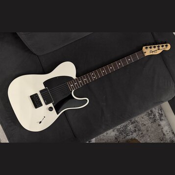 Bílá elektrická kytara s černým pickguardem a hnědým hmatníkem. Na hlavě je vidět nápis Squier by Fender a TELECASTER. Kytara má jeden snímač u kobyl…