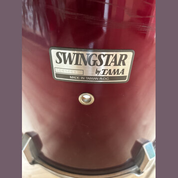 Detail červeného bubnu s nalepeným stříbrným štítkem. Na štítku je uveden nápis SWINGSTAR by TAMA a text MADE IN TAIWAN R.O.C. V levé části štítku je…