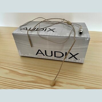 Na bílé krabici s nápisem AUDIX leží béžový hlavní mikrofon s tenkým ramínkem a konektorem. Na krabici je logo AUDIX zopakováno dvakrát. Povrch krabi…