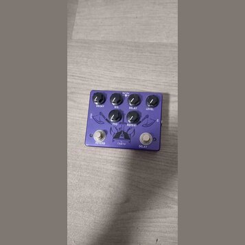 Fialový efekt pedál s kovovým povrchem. Na horní straně jsou ovládací knoflíky s popisky DECAY, MIX, DELAY, LEVEL, PRD a REPEAT. Ve spodní části jsou…