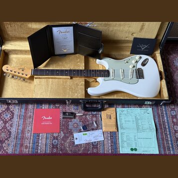 Bílá elektrická kytara typu Stratocaster s bílým pickguardem, umístěná v černém kufru. Na hlavě kytary je viditelné logo Fender. K nástroji patří cer…