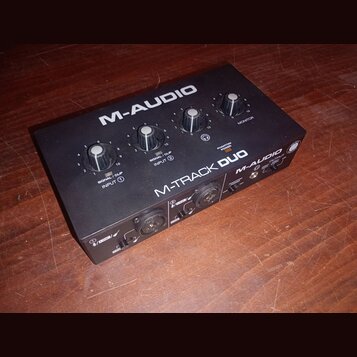 Černý audio rozhraní s nápisy M-AUDIO a M-TRACK DUO. Na horní straně jsou čtyři ovládací knofíky a kontrolky s popisky SIGNAL / CLIP INPUT 1, SIGNAL…