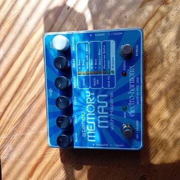 Modrý efekt pedál s bílým ovládacím knoflíkem a čtyřmi černými knoflíky. Na povrchu je nápis stereo MEMORY MAN with Hazarai a značka electro-harmonix…