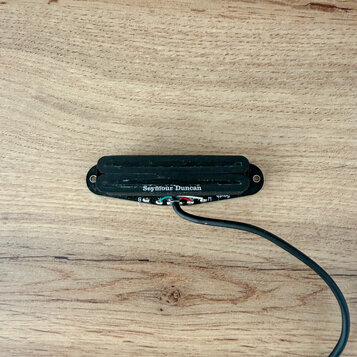 Černý humbucker s krycí maskou, umístěný na dřevěném povrchu. Na snímači je bílý nápis Seymour Duncan. Z spodní části vedou připojovací vodiče v čern…