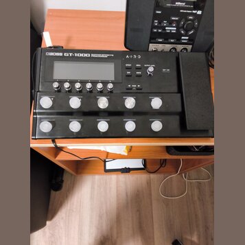 Černý kytarový procesor s nápisem BOSS GT-1000 GUITAR EFFECTS PROCESSOR a logem A.I.R.D. Zařízení má displej, pět malých tlačítek s čísly 1 až 5 a de…