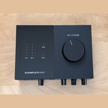 Černý audio rozhraní s matným povrchem. Na horní části jsou nápisy OUT 1/2 VOLUME, USB, 48 V a označení vstupů IN 1 a IN 2 u sloupců s kontrolkami. V…