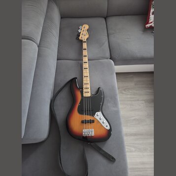 Elektrická basová kytara s korpusem v barevném přechodu od žluté přes oranžovou k červené a černé. Nástroj má černý pickguard, stříbrný most a ladící…