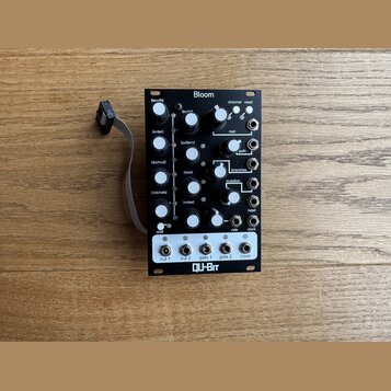 Černý modulární syntetizátor s bílými popisky a ovládacími prvky. Na horní části je nápis Bloom, v dolní části bílý panel s logem QU-Bit a výstupy oz…