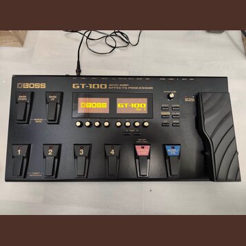 Černý kytarový procesor s nápisem BOSS GT-100 COSM AMP EFFECTS PROCESSOR. Zařízení má displej s oranžovým textem BOSS GT-100 Ver. 2, několik ovládací…