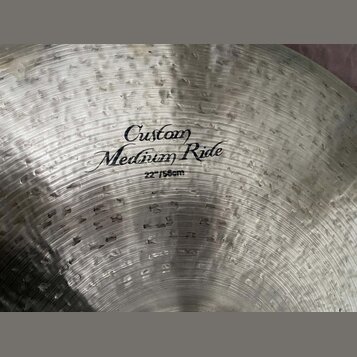 Detailní pohled na kovový činel s viditelným kovaným povrchem. Uprostřed je černý nápis „Custom Medium Ride“ a pod ním označení „22"/56cm“. Povrch vy…