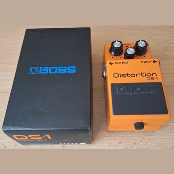 Kytarový efekt v oranžovém kovovém provedení s černým pedálem. Na zařízení jsou nápisy Distortion, DS-1 a logo BOSS. Vedle efektu je černá krabice s…